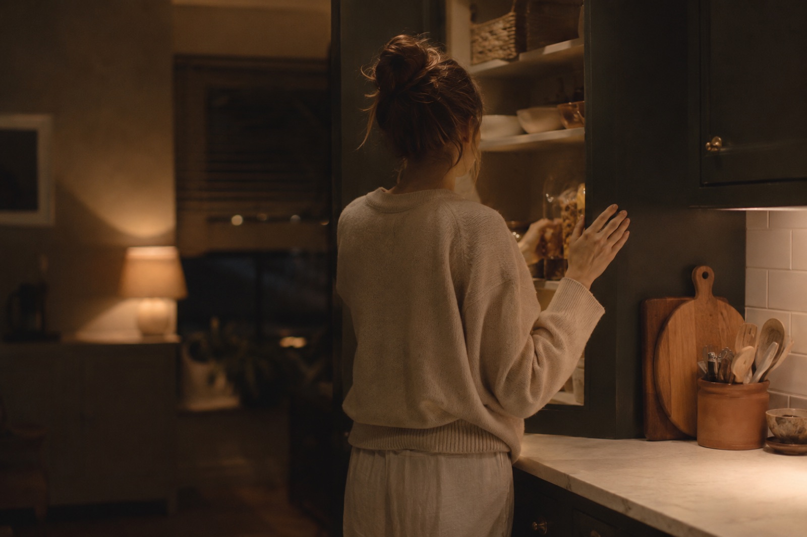 A person standing at an open kitchen cupboard in the evening, snacking out of habit rather than hunger.